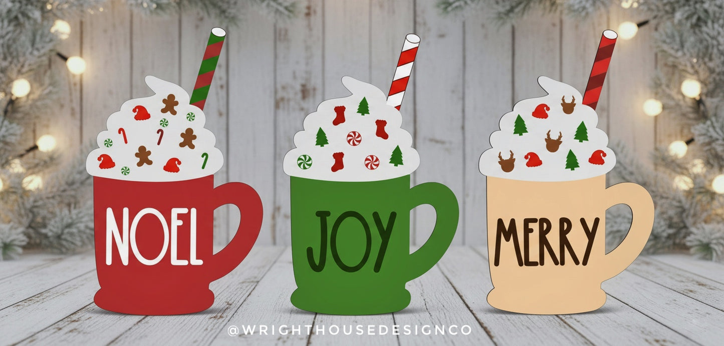 Three festive mugs with 'NOEL', 'JOY', and 'MERRY' text on a wooden surface with Christmas lights in the background.